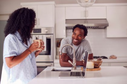 Couple Wearing Pyjamas Standing In Kitchen Working From Home On Laptop